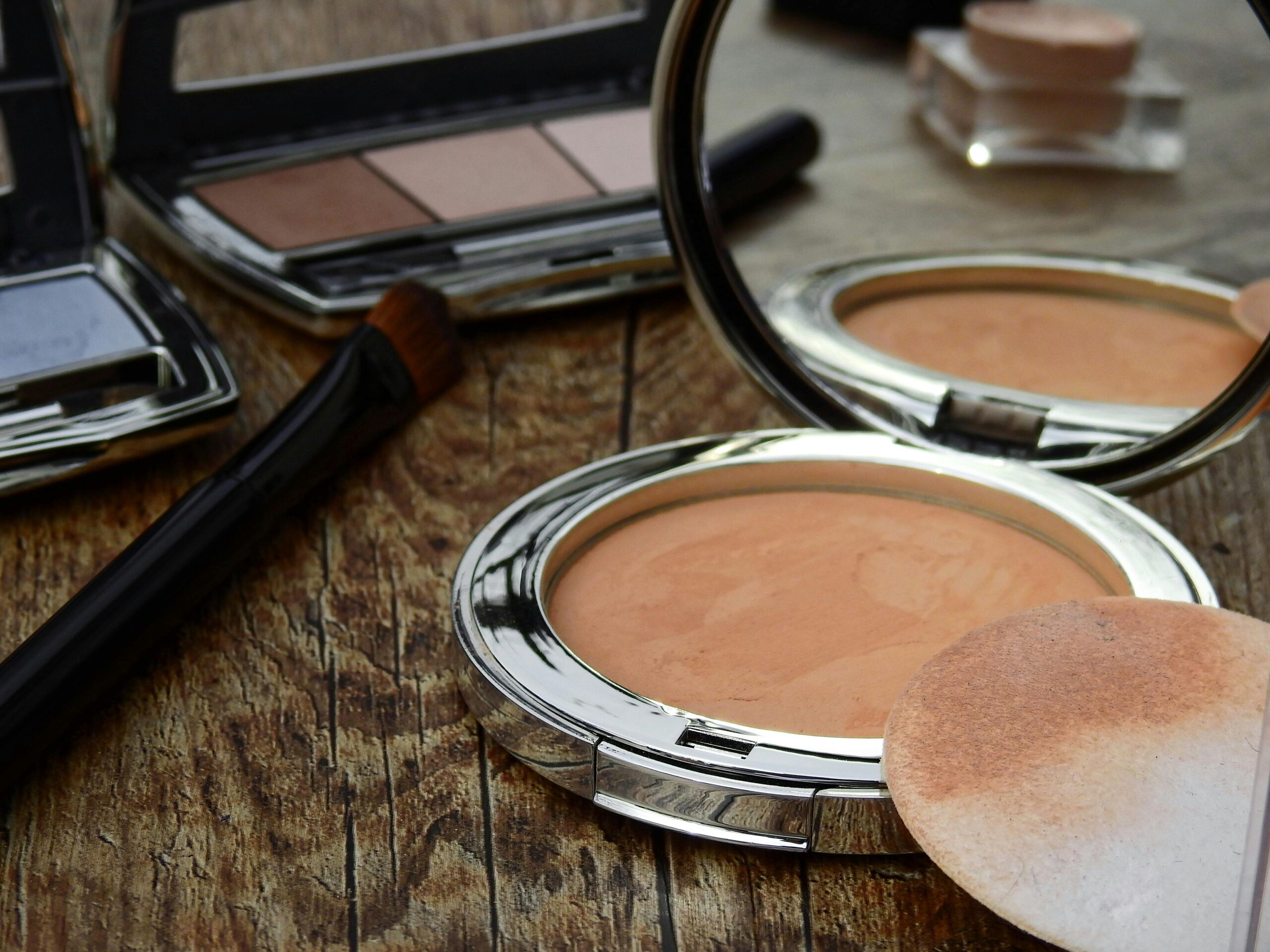 Close-up of makeup tools including compact, brush, and mirror on a rustic wooden surface.